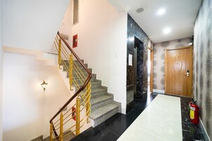 Uma escadaria com guarda-corpo de madeira e balaústres amarelos, que conduz a um quarto com chão de mármore e uma porta com placa de número.