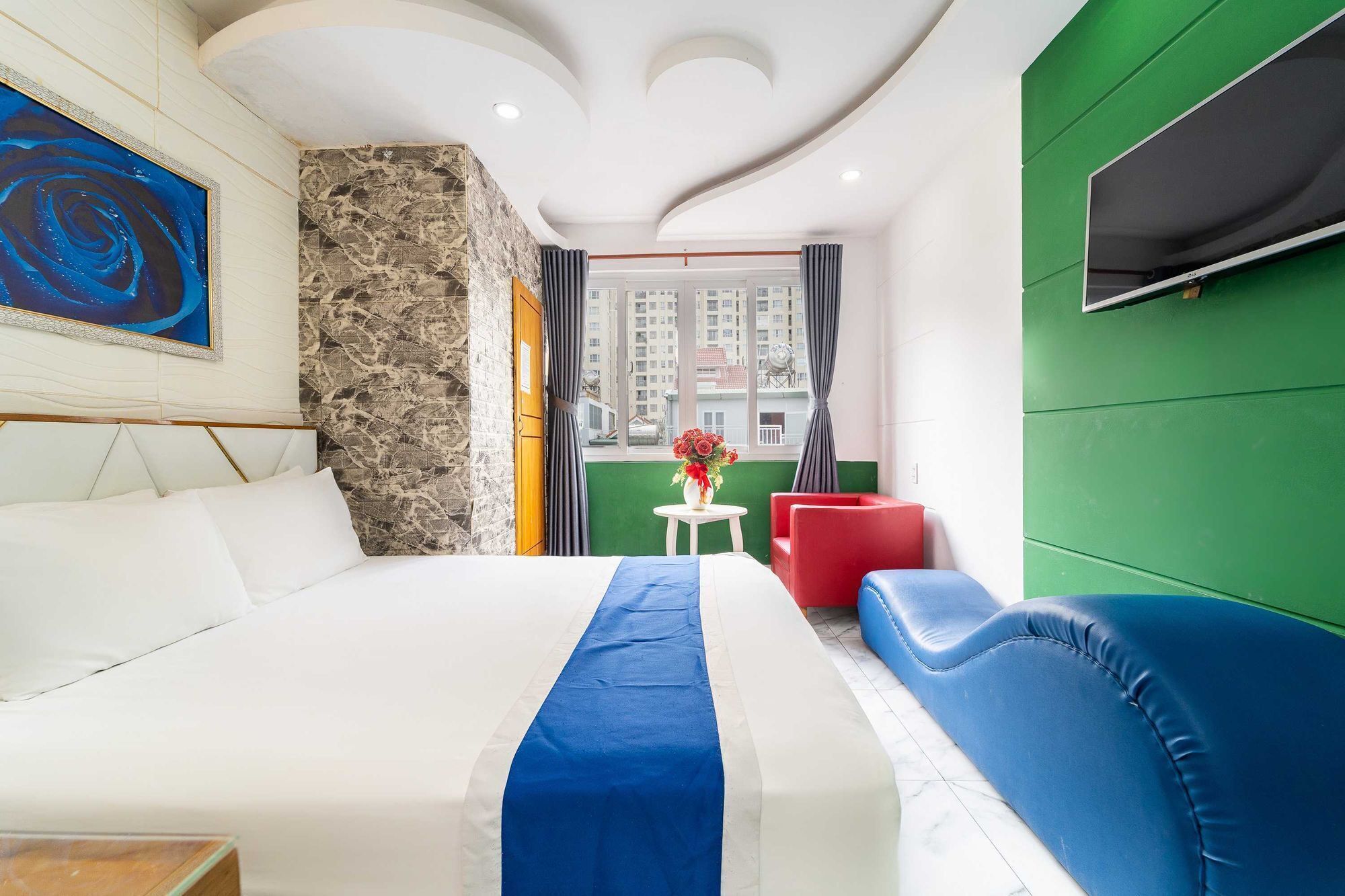 A hotel room with a large bed, a red chair, a blue sofa, a stone accent wall, a painting, and a TV on a green panel.