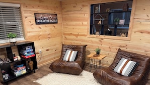 A cozy cabin-style room with two leather sofas, a small wooden side table, and a shelf with board games. A sign on the wall reads "Welcome to our NECK OF THE WOODS."