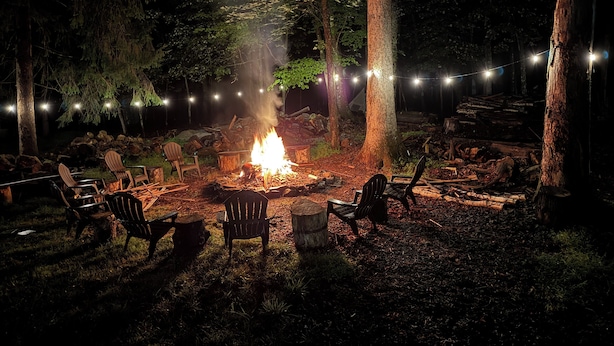 A campfire surrounded by chairs in a forested area with string lights.