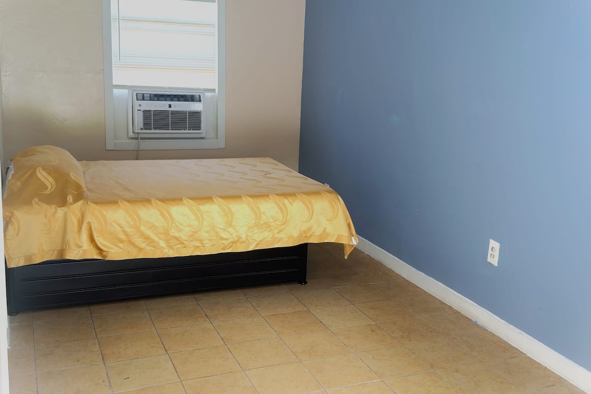 A small, single-bed room with a window, air conditioning unit, and tiled floor.