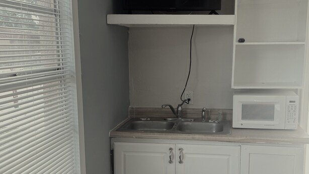 A small kitchenette with white cabinets, a microwave, a sink, and a window with blinds. - Star Motel (Tampa)