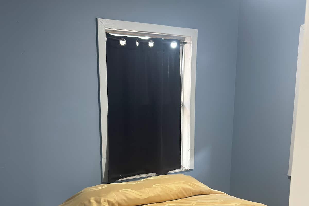 A single bed with a patterned bedspread in a room with light blue walls and a window with a black curtain.