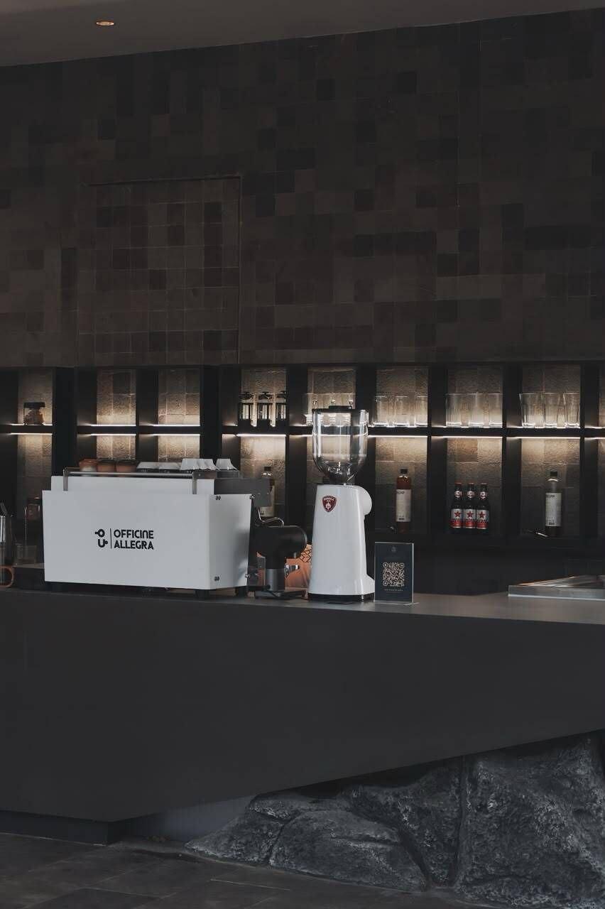 Zona bar moderna con una macchina per caffè e un frullatore, scaffali con bicchieri e un codice QR per interazioni digitali.