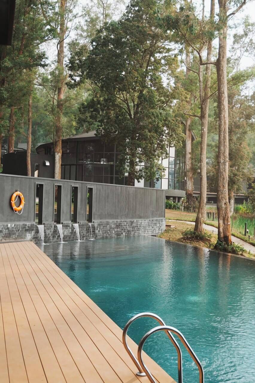 A swimming pool with a wooden deck, a waterfall feature, and a modern building with glass windows.