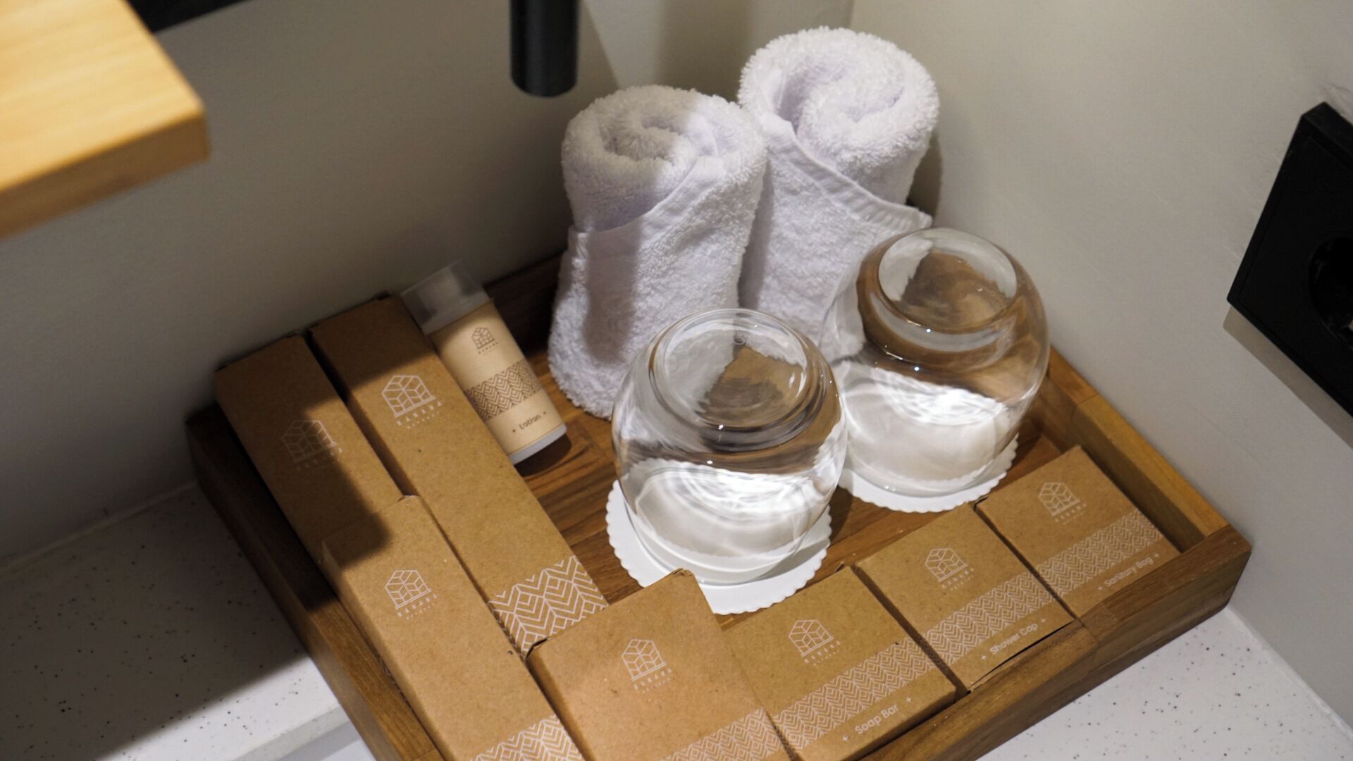 Bandeja de madera con artículos de aseo, incluyendo una pastilla de jabón, gorro de ducha y dos botellas de vidrio, presumiblemente con loción o champú, junto a toallas blancas dobladas con esmero.