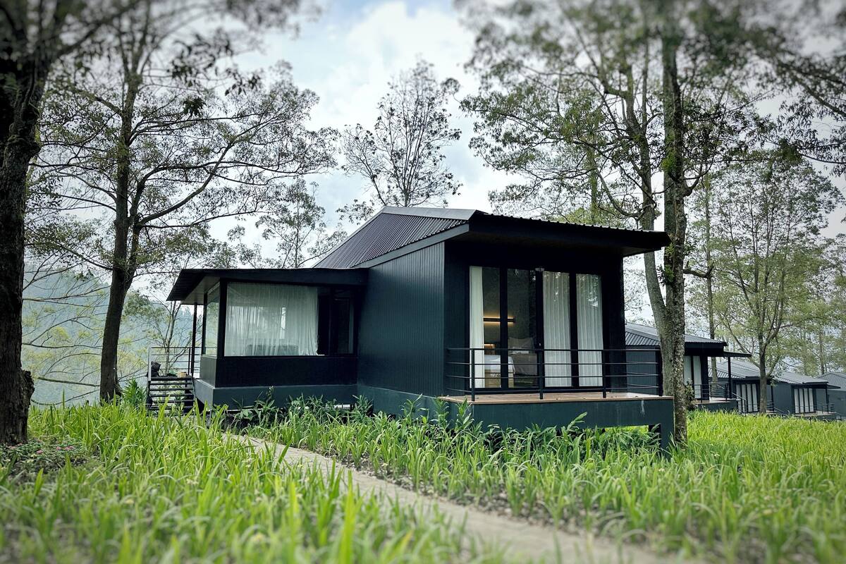 A modern, dark-colored cabin with a glass balcony, surrounded by lush greenery and trees.