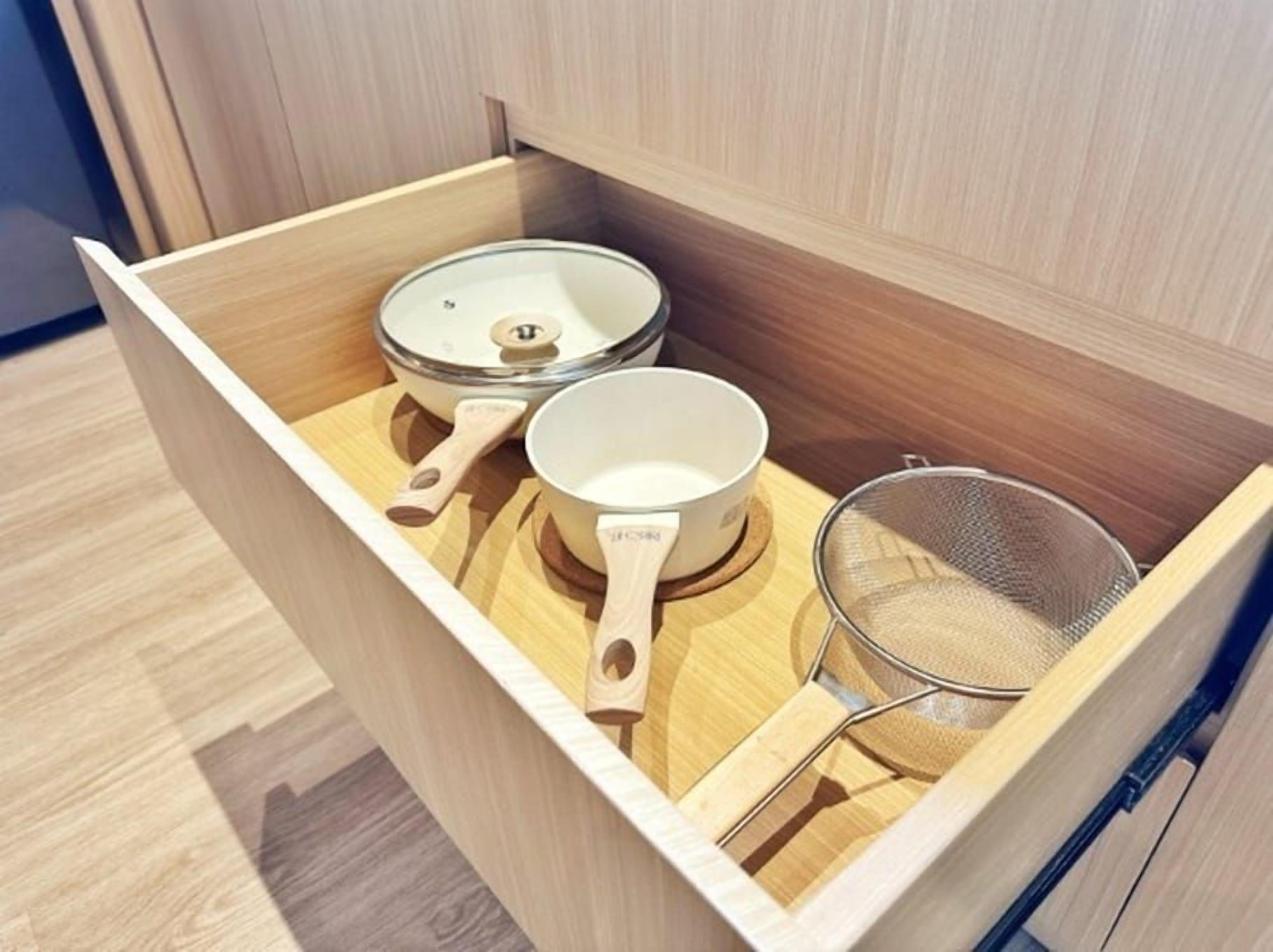 A wooden drawer with kitchen utensils including a strainer and two pots.