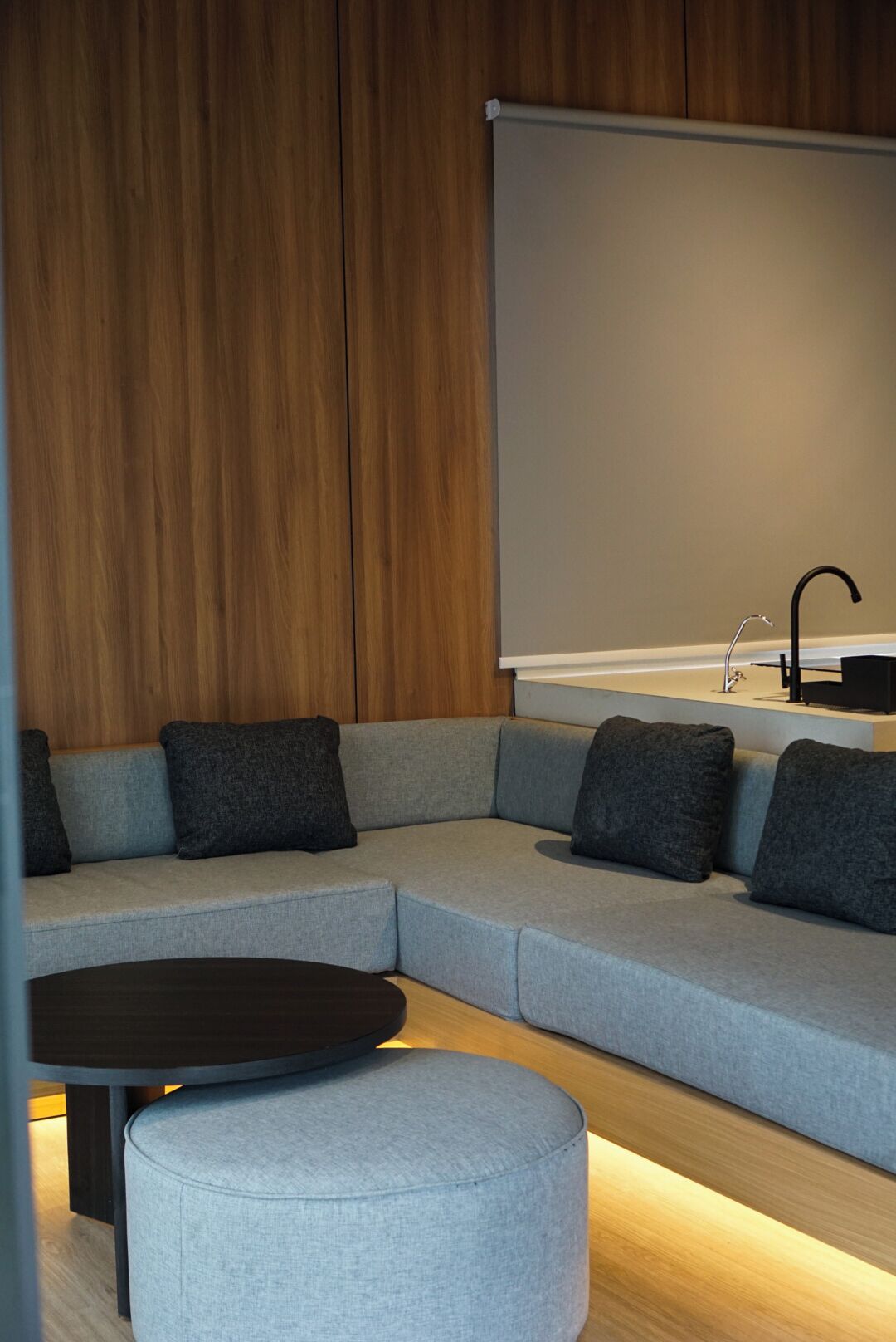A modern living room with a grey sectional sofa, dark grey cushions, a round ottoman, a black coffee table, a wooden accent wall, and a large blank projection screen.