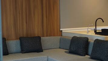 A modern living room with a grey sectional sofa, dark grey cushions, a round ottoman, a black coffee table, a wooden accent wall, and a large blank projection screen.