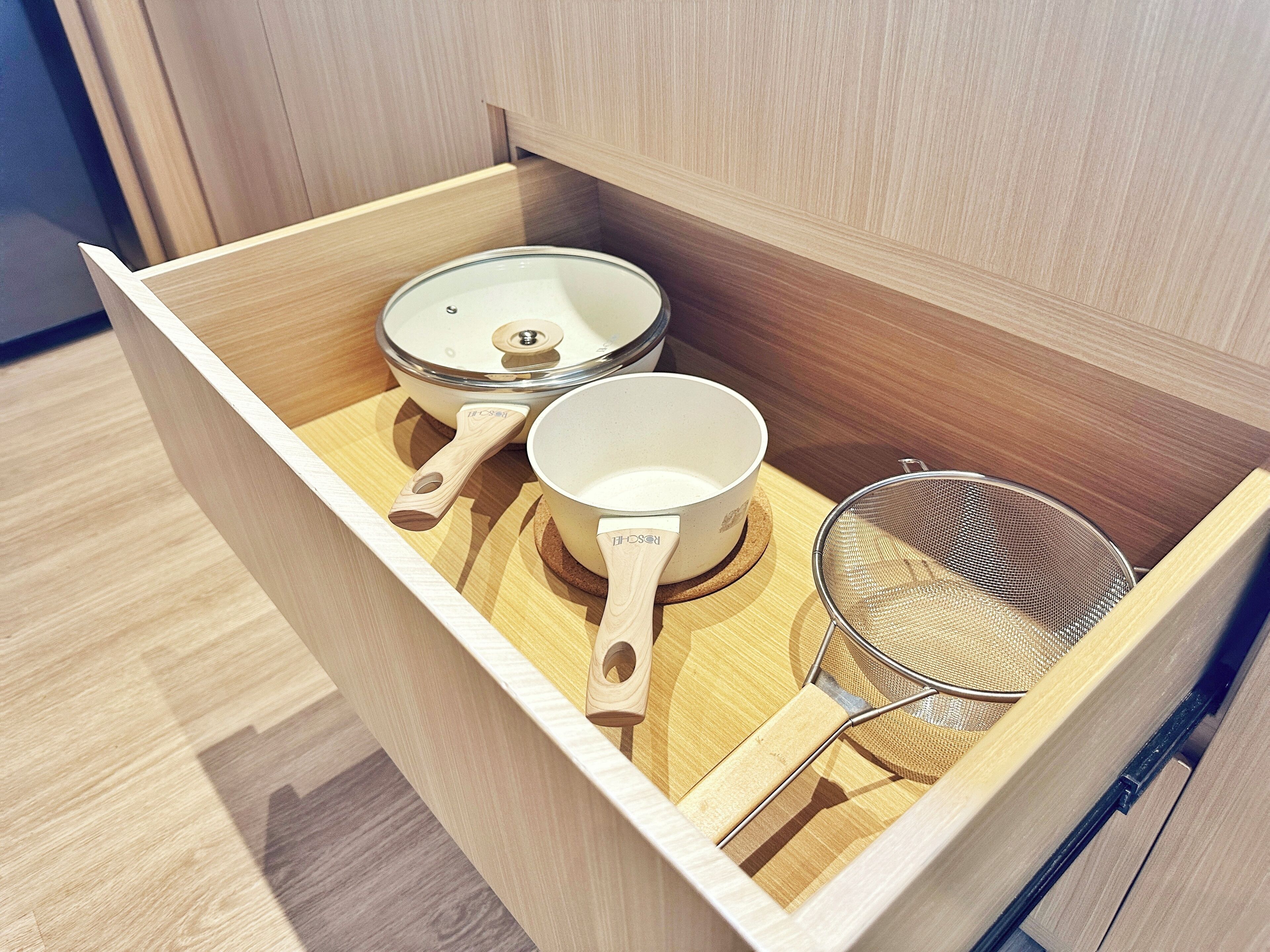 A wooden drawer containing kitchen utensils including a strainer, a pot, and a pan.