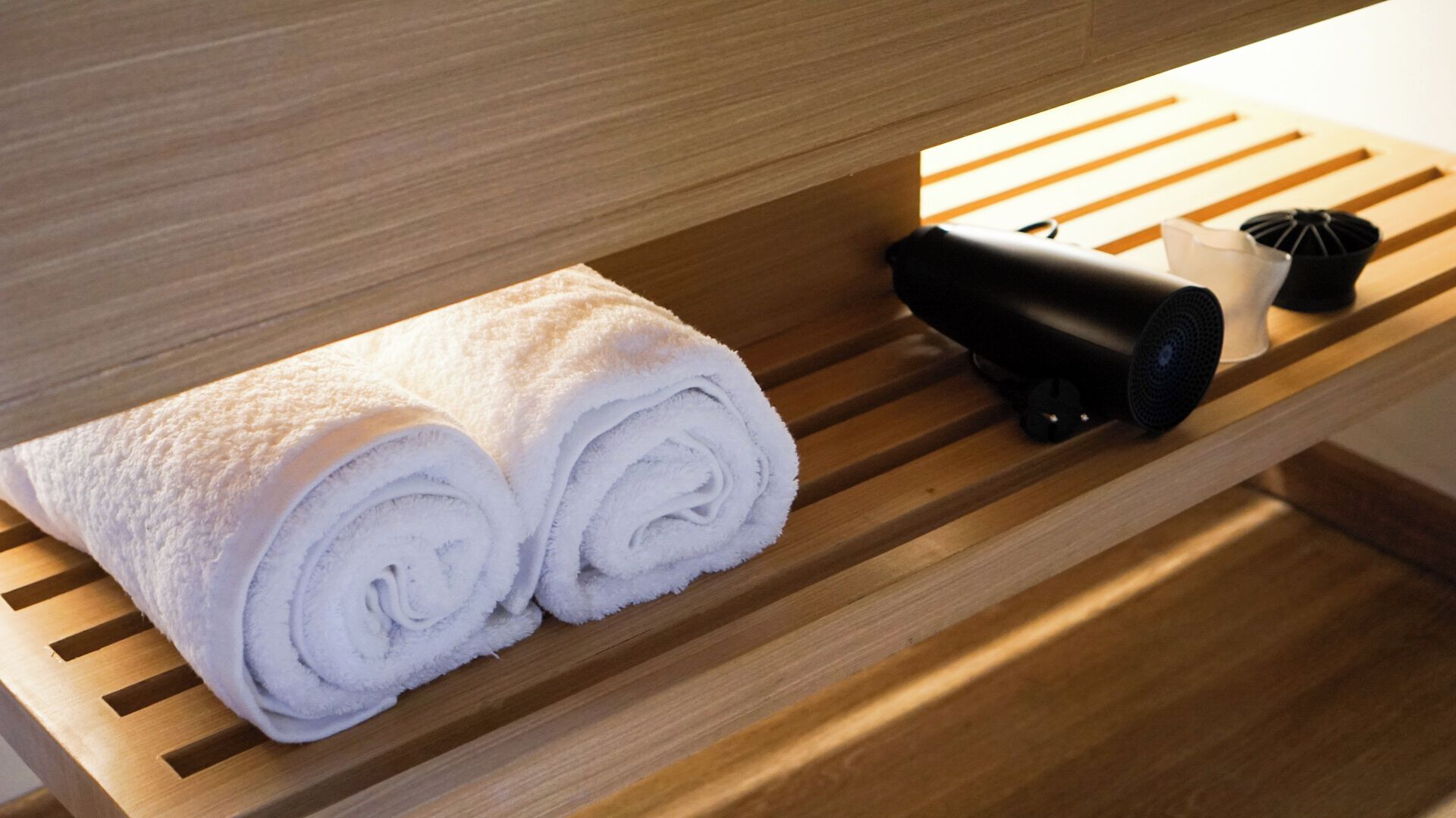 A wooden bench with two rolled white towels and a black hairdryer.