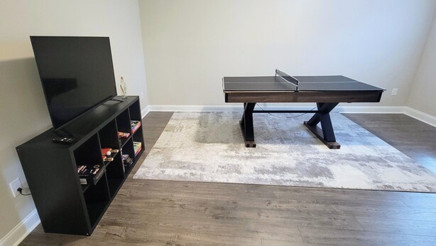 Une pièce avec un téléviseur à écran plat posé sur un meuble noir, une table de ping-pong et un tapis blanc.