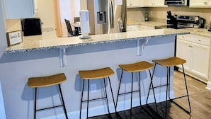 Une cuisine moderne dotée d’un îlot central, de tabourets de bar, d’appareils électroménagers en acier inoxydable et de meubles de rangement blancs.
