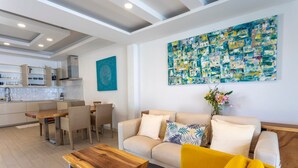 Un salon moderne avec un canapé jaune, une table basse en bois et un divan beige. On aperçoit à l’arrière-plan un coin cuisine avec des meubles blancs et une table à manger.