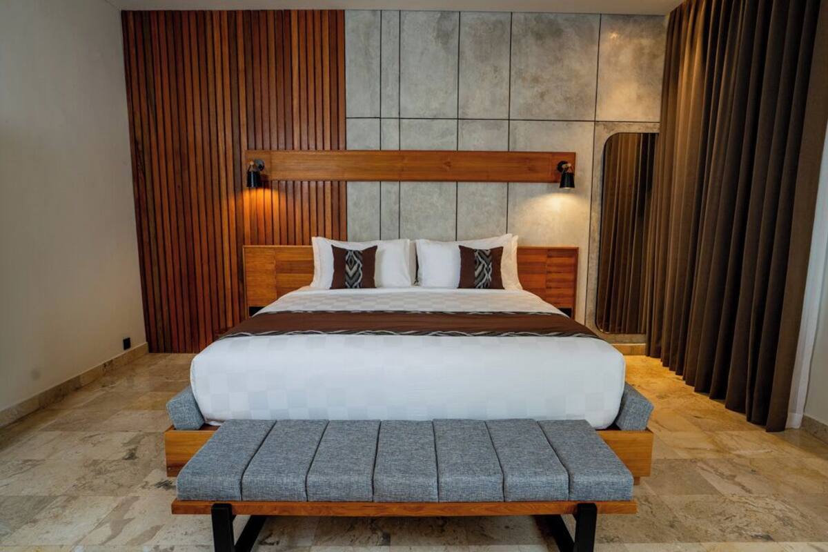 A modern bedroom with a wooden headboard, a large bed with grey and white bedding, and a bench at the foot of the bed.
