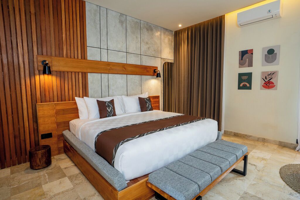 Une chambre à coucher moderne avec une tête de lit en bois, un grand lit agrémenté d’un banc gris et un mur en pierre orné d’œuvres d’art.