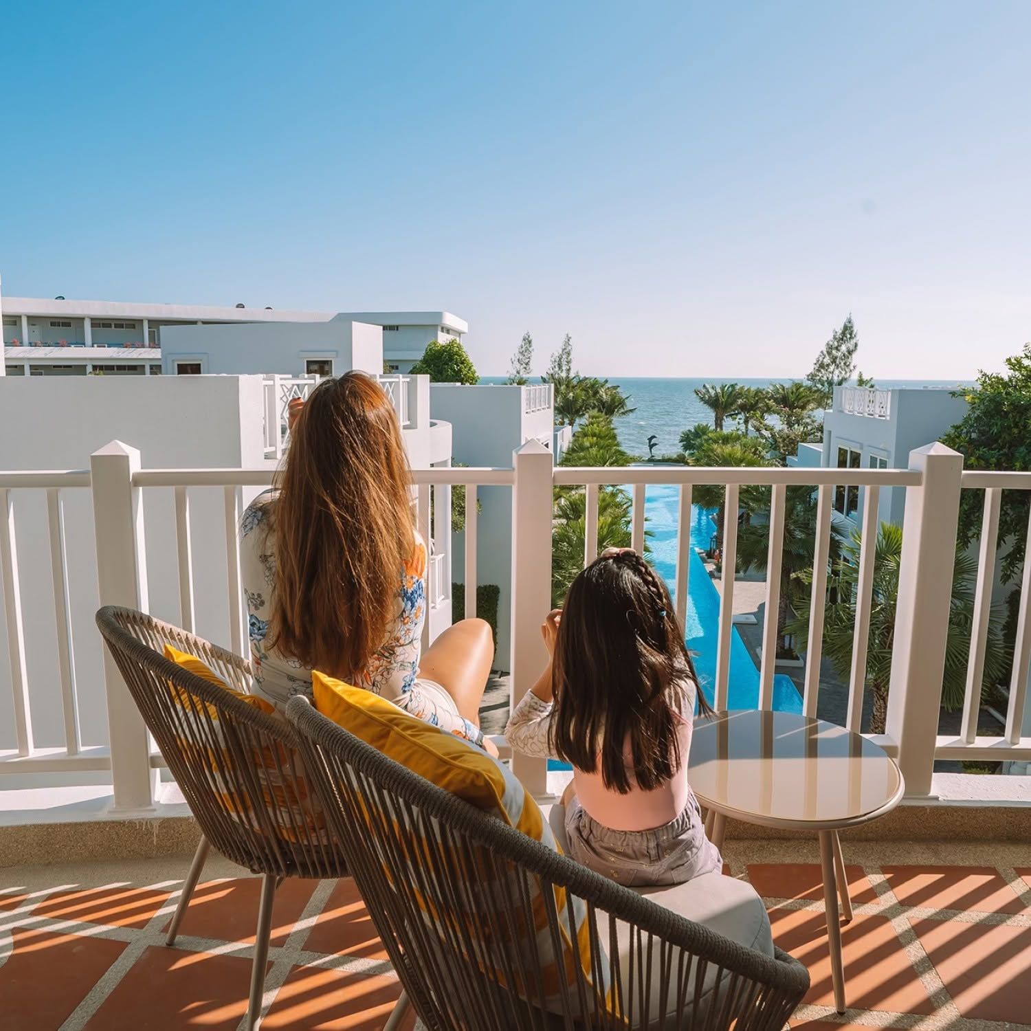 Two people seated on a balcony overlooking a swimming pool and the sea.