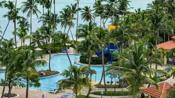 A resort with a pool surrounded by palm trees and a view of the ocean. - Rooftop Terrace Condo with Golf Views – 5-Min Walk to Palmas del Mar Beach FL671 (Humacao)
