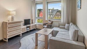 Ein modernes Wohnzimmer mit einer Couch, einem Fernseher und einem kleinen Tisch. Große Fenster mit Vorhängen bieten Blick auf Häuser im Freien.