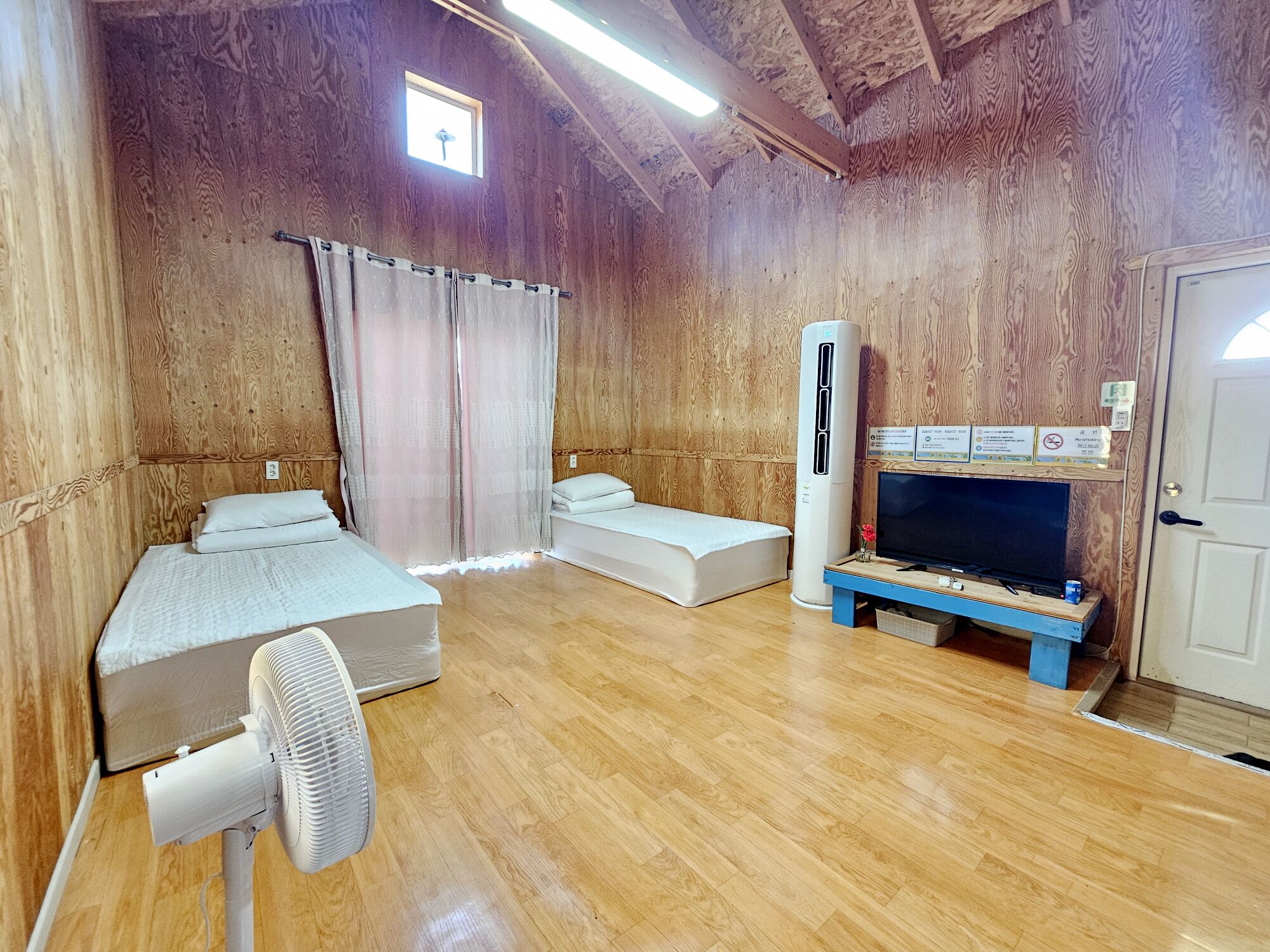 Une pièce aux murs en bois, avec un ventilateur de plafond, deux lits, un téléviseur et une petite fenêtre.