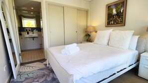 A bedroom with a bed, two bedside tables, a nightstand, a painting, and a bathroom visible through an open door. - Sunlit & Peaceful Contemporary Apartment (Randwick)