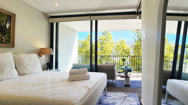 A hotel room with a large bed, a chair, a small table, and a balcony with a view of trees and a building. - Sunlit & Peaceful Contemporary Apartment (Randwick)