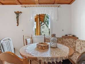 A cozy dining area with a round wooden table, lace tablecloth, and a rustic wooden centerpiece. A wooden chair is visible, and a window with a view of a house and trees is in the background.