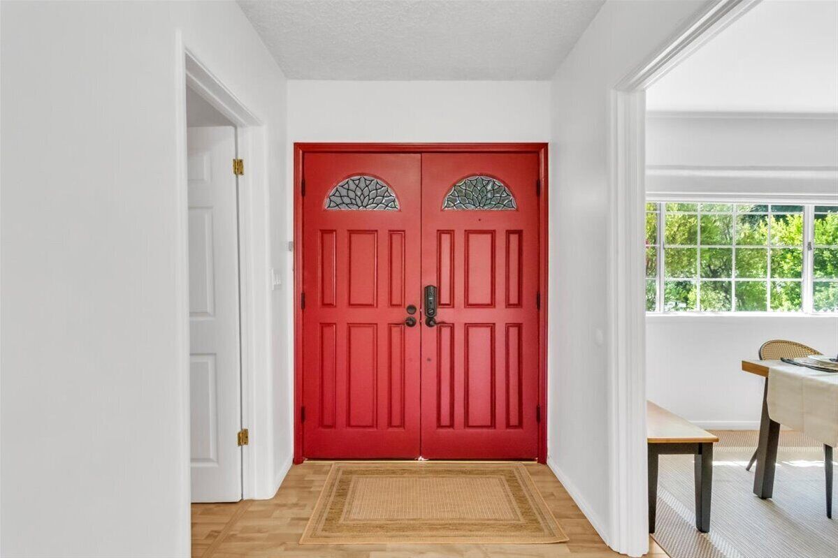 Una puerta roja con paneles de vidrio, suelo de madera y ventana con vista a la vegetación.