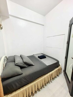 A single bed with a dark mattress and multiple pillows, a bedside table, and a small window with blinds. - COZY MINIMALIST HOME NEAR NAIA TERMINALS PARANAQUE (Parañaque)