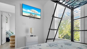 Une chambre à coucher avec un lit, un téléviseur diffusant une scène de plage, une échelle et une fenêtre avec des stores.