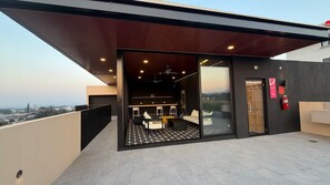 Een moderne, overdekte lounge met een glazen deur die leidt naar de binnenruimte, een plafondventilator en een brandblusser.