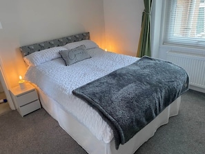 A neatly made bed with a grey blanket, a pillow with text, and a bedside lamp.