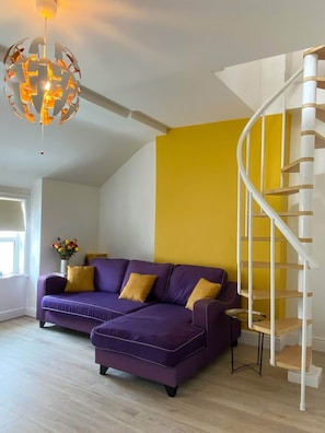 A modern living room with a purple sofa, a yellow accent wall, a white spiral staircase, and a unique spherical light fixture.
