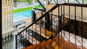 A staircase with a wooden handrail and black metal balusters, leading to a balcony with a view of a swimming pool and greenery.