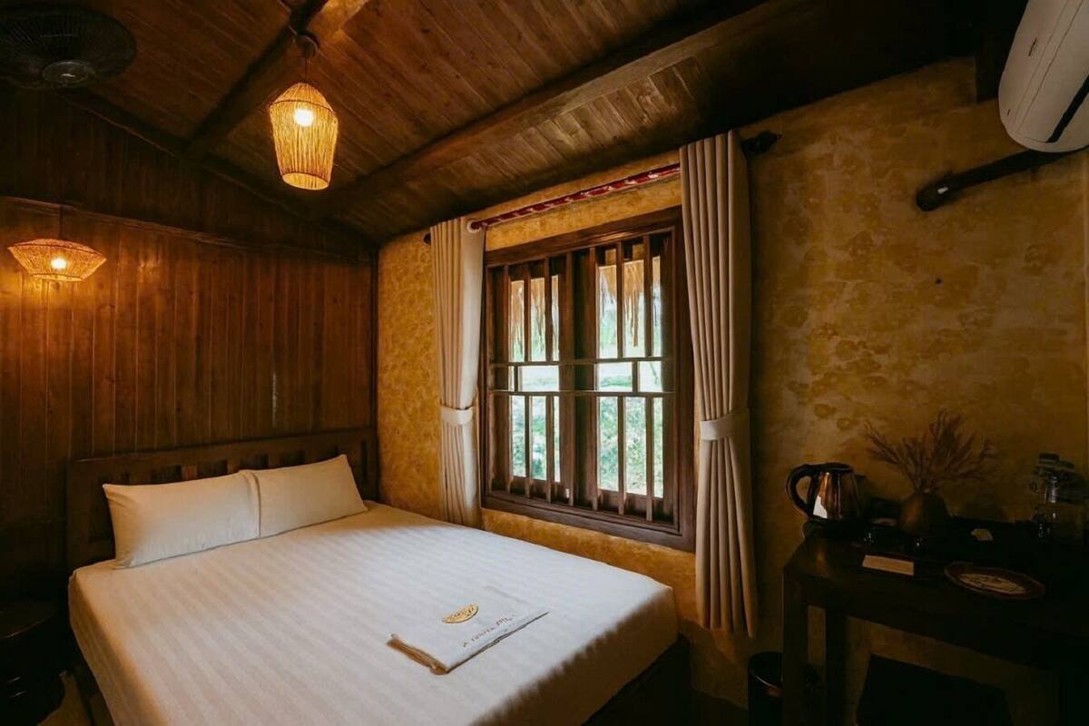 A wooden room with a bed, a window with curtains, a small table with a kettle, and a wall-mounted heater.