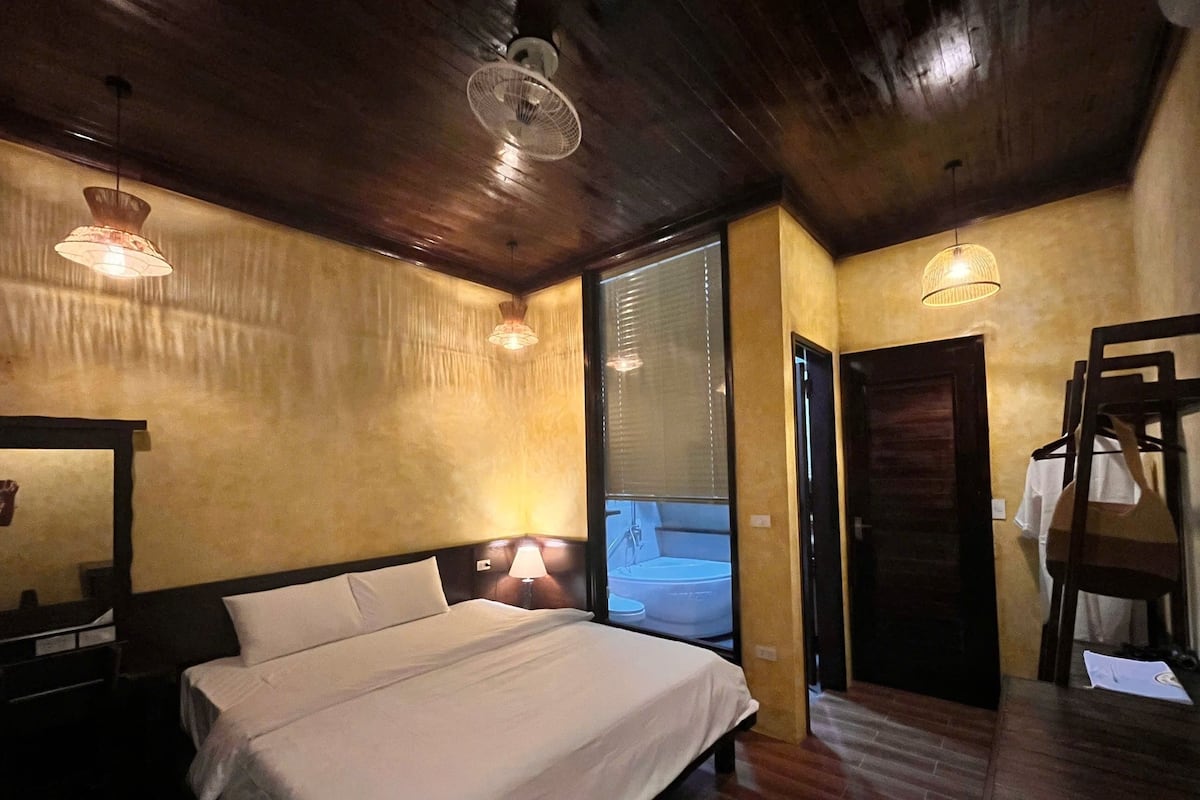 A bedroom with a large bed, wooden ceiling, and a window with blinds.