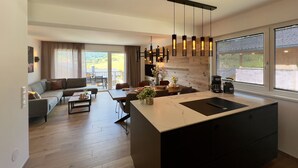 A modern kitchen with a central island, a dining area with a sofa and coffee table, and a view of the outdoors through large windows.