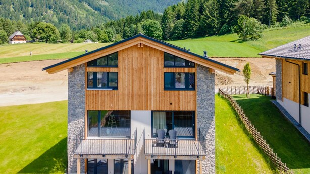 A two-story wooden house with a balcony, surrounded by a fenced property and a mountainous landscape.
