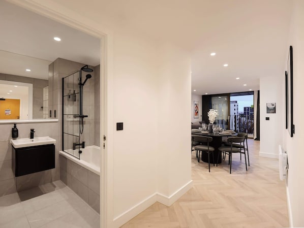 A modern bathroom with a shower, sink, and toilet. There is a dining area with a table and chairs, and a view of the city through large windows.