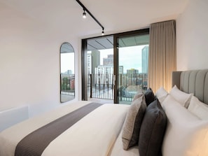A modern hotel room with a large bed, a view of the city through floor-to-ceiling windows, and a balcony with a railing.