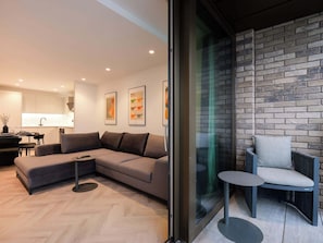 A modern living room with a sectional sofa, a small round table, and a glass partition with a brick wall. - Solaro | The Heim (Greater Manchester)