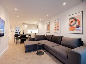 Una sala de estar moderna con un sofá modular, un comedor con mesa y sillas, una cocina al fondo y obras de arte en las paredes.
