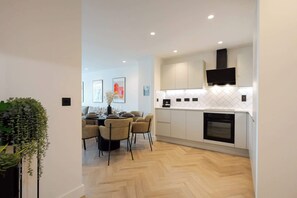A modern kitchen with white cabinets, a built-in oven, and a dining area with a table set for four. - Urbano | The Heim (Greater Manchester)