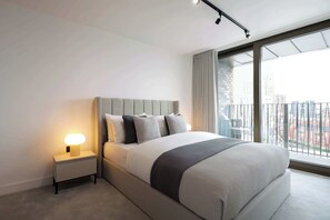 A modern hotel room with a large bed, a bedside table with a lamp, and a view of the city through large windows.