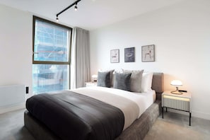 A modern bedroom with a large bed, a nightstand, a lamp, and a window with a view of a building. - Urbano | The Heim (Greater Manchester)