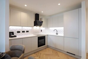 A modern kitchen with white cabinetry, a black range hood, a built-in oven, and a herringbone-patterned backsplash. - Urbano | The Heim (Greater Manchester)