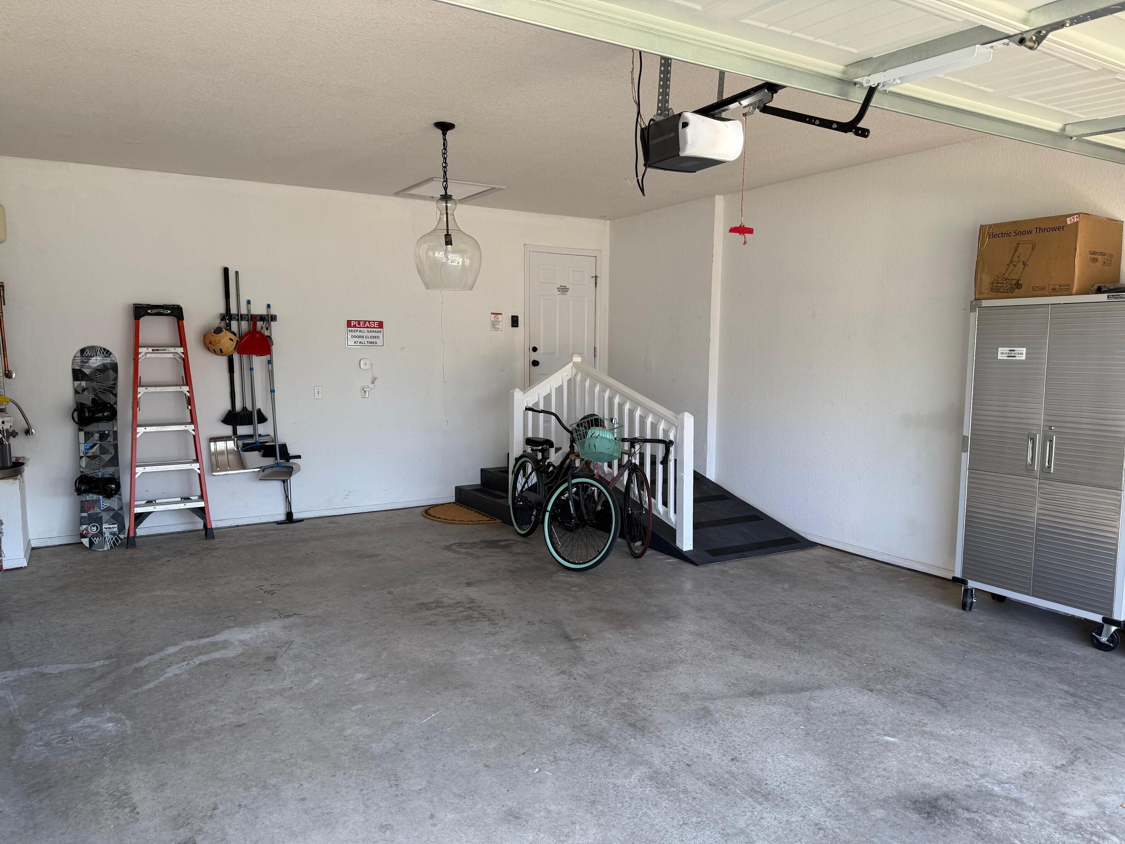 Un garage équipé d’un ventilateur de plafond, d’un projecteur fixé au plafond, d’un vélo avec un panier, d’un coffre pour un déneigeuse et d’une armoire métallique.