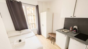 A compact hotel room with a bed, a small kitchenette, and a view of the city.