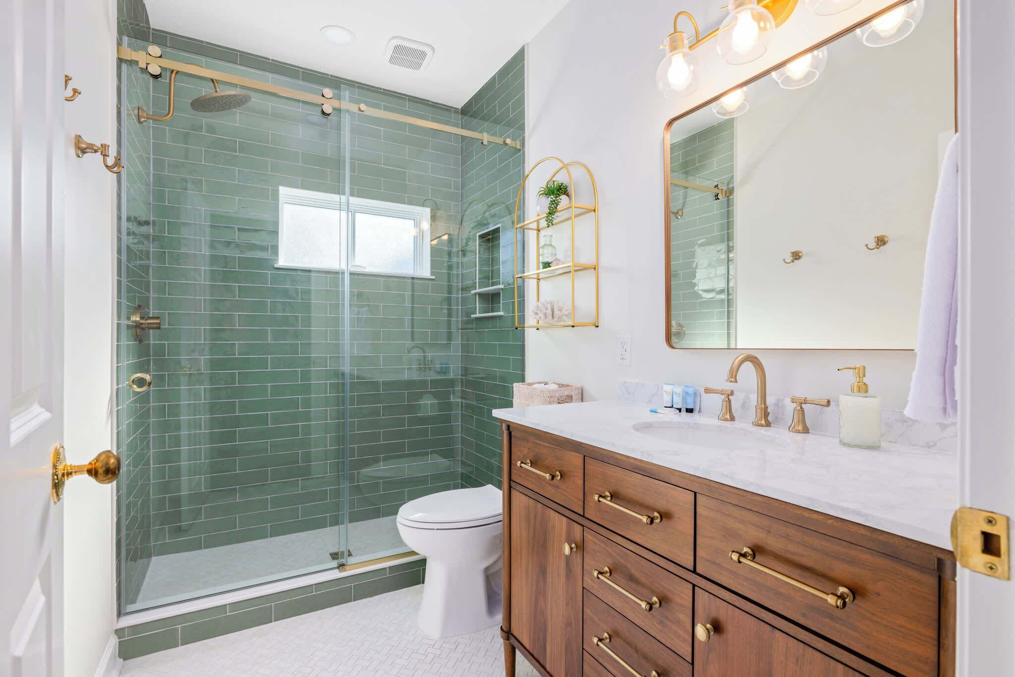 A bathroom with a glass shower enclosure, a white toilet, a vanity with gold fixtures, and a mirror with lights.